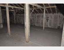 Dachboden eines alten Gebäudes mit freiliegenden Holzbalken, einem Holzfußboden und unverkleideten Wänden aus brüchigem Material; die Konstruktion zeigt sichtbare Abnutzung und Spuren von Verwitterung.