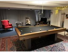 Ein Hobbyraum mit einem Billardtisch in der Mitte, umgeben von Sesseln, darunter ein roter Polstersessel und ein Stuhl mit einer Plüschfigur. Im Hintergrund befinden sich Vorhänge, eine kleine Sitzecke mit Büchern und eine Wand mit Billardqueues. Der Raum ist mit Orientteppichen ausgelegt und durch warmes Licht beleuchtet.