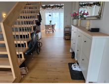 Heller Eingangs- und Flurbereich eines Wohnhauses mit Holztreppe links und einem weißen Sideboard mit großem Wandspiegel rechts. Im Hintergrund führt der Raum zum Essbereich mit Tisch vor einer großen Glas-Schiebetür; Holzfußboden und Blumen-/Girlandendekoration sind sichtbar.