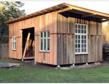 Ein einfaches Holzgebäude mit vertikaler Paneelstruktur und großen rechteckigen Fenstern, umgeben von Gras und Bäumen. Ein offener Eingang zeigt den Innenraum mit Holzbalken und Bauwerkzeugen.