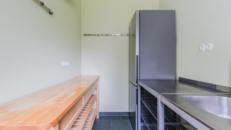 Schmaler Küchen- oder Hauswirtschaftsraum mit hellgrünen Wänden, grauen Fliesen und Edelstahl-Arbeitsfläche mit Spülbecken. An der Seite stehen ein hoher Kühlschrank und offene Metallregale, gegenüber eine lange Holzablage mit Untergestell sowie Wandhaken und Steckdosen.