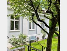 Weiße Fassade eines Wohnhauses mit großen Fenstern und einem gepflegten Garten im Vordergrund, in dem ein Baum mit dichtem Laub sowie eine hellblaue Holzbank vor der Hauswand stehen.