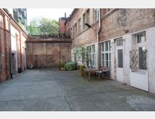 Ein rechteckiger Hinterhof mit Backsteinfassaden, großen Fenstern, weißen Türen und einem Betonboden. Der Hof ist spärlich ausgestattet mit einem Tisch, Stühlen und Pflanzen in Töpfen; im Hintergrund steht eine hochgezogene Mauer mit einer kleinen bepflanzten Terrasse darüber.