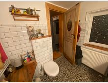 Kleines Badezimmer mit weiß gefliesten Wänden, gemustertem Bodenfliesen und einer Holztoilette. Es gibt ein Fenster mit dunkler Fensterfolie, ein Holzregal mit Dekoartikeln, und eine geöffnete Holztür führt zu einem angrenzenden Raum.