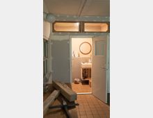 Ein Raum mit einer geöffneten Tür, die in ein einfach eingerichtetes Badezimmer mit einem runden Spiegel, einem Waschbecken und Fliesenboden führt. Im Vordergrund ein Werkraum mit Holzbalken, Werkzeugen und halbfertigen Wänden.