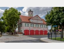 Zweistöckiges Gebäude einer städtischen Feuerwache mit cremefarbener Fassade, grünen Fensterläden und einem roten Ziegeldach. Im Erdgeschoss befinden sich fünf rote Garagentore, und ein kleiner Turm mit einer Uhr und einem Wappen überragt das Dach, umgeben von Bäumen und einer gepflasterten Straße.
