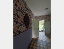 Ein Flur mit einer floralen Tapete auf der oberen Wandhälfte und hellvioletter Farbe auf der unteren, kombiniert mit einem rot-verzierten Wandornament. Der Boden ist mit braun-schwarz gemusterten Fliesen belegt, und eine geöffnete weiße Tür führt nach draußen, wo ein grüner Gartenbereich erkennbar ist.
