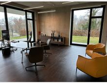 Modernes Büro mit großen Fensterfronten, die Blick auf einen Garten mit Bäumen und Hecken bieten. Der Raum ist mit einem dunklen Holzboden, einem Schreibtisch, mehreren Stühlen und gelben Sesseln eingerichtet, ergänzt durch eine Wandkonsole mit Deko-Elementen.