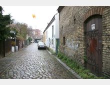 Kopfsteingepflasterte Gasse mit Backstein- und verputzten Hausfassaden, teils mit Graffiti verziert. Am Straßenrand steht ein geparktes Auto, und im Hintergrund sind Bäume und weitere Gebäude sichtbar.