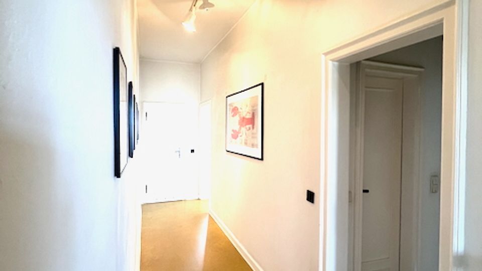 Langer, schmaler Innenflur mit weißen Wänden, glattem Boden und Deckenbeleuchtung über einer Schiene. An den Wänden hängen gerahmte Bilder, rechts öffnet sich eine Tür zu einem angrenzenden Raum mit einer weiteren weißen Tür.