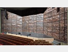 Ansicht eines Theatersaals mit Reihen von Polstersitzen im Vordergrund und einer Bühne mit Holzverkleidung im Hintergrund. Die Rückwand der Bühne ist mit einer kunstvoll strukturierten, reliefartigen Oberfläche in warmen Tönen gestaltet.