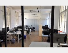 Großraumbüro mit mehreren Schreibtischen, Bürostühlen und Computern, helle Wände, große Fenster mit Lamellen und Deckenventilatoren. Eine Person arbeitet an einem Schreibtisch im Hintergrund.