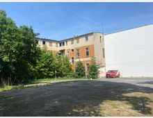 Ein leerer Parkplatz vor einem teilweise verfallenen Gebäude mit gemauerten und verputzten Außenwänden, bewachsen von Bäumen und Sträuchern. Rechts steht ein rotes Kleintransporterfahrzeug, der Hintergrund zeigt einen klaren blauen Himmel.