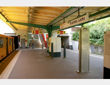 U-Bahn-Station mit gelbem Zug auf der linken Seite, überdachtem Bahnsteig und einem Schild mit der Aufschrift „Krumme Lanke“. Im Hintergrund führen Treppen nach oben, und eine grüne Umgebung ist rechts vom Bahnsteig sichtbar.