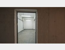 Blick in einen leeren Raum mit weiß gestrichenen Betonwänden, einem grauen Betonboden und rechteckigen Deckenleuchten. Der Eingang ist von gemauerten Wänden umrahmt, die teilweise beige gestrichen sind.