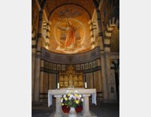 Innenraum einer Kirche mit einem geschmückten Altar im Vordergrund. Über dem Altar befindet sich ein goldenes Mosaik, das Christus mit ausgebreiteten Armen darstellt, umgeben von detaillierten, farbigen Verzierungen und Arkaden aus Marmor.
