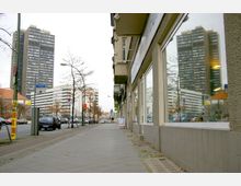 Straßenszene mit Gehweg, auf dem rechts eine Fensterfront eines Gebäudes und links eine Straße mit parkenden Autos zu sehen sind. Im Hintergrund befinden sich moderne Hochhäuser und Bäume ohne Laub.