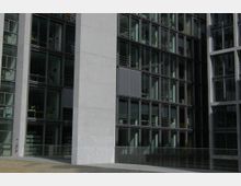 Außenansicht eines modernen Bürogebäudes mit einer Fassade aus großen Glasfenstern in rechteckigen Metallrahmen. Dazwischen befinden sich Betonpfeiler, und einige Fenster zeigen Einsichten in Büros mit Regalen und Schreibtischen.