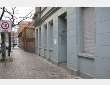 Straßenszene in einer Wohngegend mit einer grauen Gebäudefassade und kleinen Fenstern; ein Schild mit Geschwindigkeitsbegrenzung auf 10 km/h steht links, daneben eine gepflasterte Gehwegfläche und kahlen Bäumen. Weiter hinten ist eine rote Ziegelmauer und eine weitere Reihe von Gebäuden sichtbar.