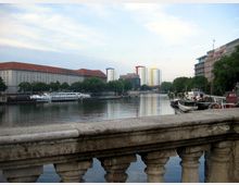 Blick von einer Steinbalustrade auf einen Fluss, gesäumt von historischen Gebäuden mit roten Dächern auf der linken Seite und modernen Wohnhäusern sowie Booten auf der rechten Seite. Im Hintergrund sind mehrstöckige, bunt gestaltete Hochhäuser sichtbar.