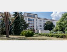 Ein mehrstöckiges Gebäude mit heller Putzfassade und großen Fenstern, umgeben von hohen Bäumen und einer Grünfläche im Vordergrund. Im Gartenbereich steht ein kleines, weißes Gewächshaus.