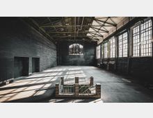 Große, leere Industriehalle mit dunkelgrauen Backsteinwänden, einer hohen Decke mit sichtbaren Metallträgern und großen, vergitterten Fenstern, durch die Licht einfällt. Im Vordergrund steht ein kleines Backsteingeländer um eine Treppenöffnung.