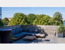 Holzterrasse mit einer grauen Eck-Couch, dekoriert mit kleinen Tischen und Pflanzen. Im Hintergrund sind Bäume und ein blauer Himmel zu sehen, mit roten Dachhäusern und einem Fluss am rechten Bildrand.