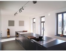 Moderne Küche mit einer großen Kücheninsel in dunklem Grau, integriertem Waschbecken und Arbeitsflächen aus Edelstahl. Der Raum ist hell, mit weißen Wänden, hohen Fenstern und schlichten, geometrischen Wanddekorationen.