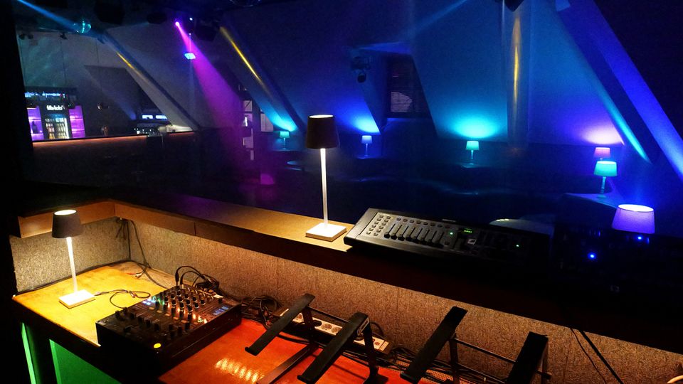 Innenansicht eines Nachtclubs mit DJ-/Technikpult im Vordergrund, Mischpult und Kabeln sowie kleinen Tischlampen entlang einer niedrigen Trennwand. Im Hintergrund sind Lounge-Sitzbereiche und eine Bar zu sehen, beleuchtet von farbigen Scheinwerfern und LED-Licht in Blau, Violett und Pink unter einer schrägen Decke.