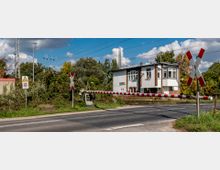 Bahnübergang mit geschlossener Schranke und rotem Warnschild, im Hintergrund ein weißes zweistöckiges Gebäude und Bäume. Über den Gleisen verlaufen Oberleitungen, die Umgebung ist von Vegetation umgeben.