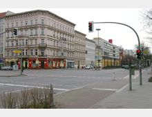 Straßenecke in einer Stadt mit mehrstöckigen Altbaugebäuden und Geschäften im Erdgeschoss, darunter eine Kneipe. Im Vordergrund eine Kreuzung mit Verkehrsampeln und Zebrastreifen, im Hintergrund moderne Bürogebäude und eine U-Bahn-Station.