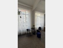 Heller Innenraum mit hohen Sprossenfenstern und bodenlangen, transparenten Vorhängen sowie sichtbaren Deckenbalken und einer weißen Backsteinwand. Vor den Fenstern steht eine kleine runde Tischgruppe mit schwarzen Stühlen und einem blauen Hocker auf einem hellen Teppich.