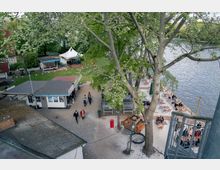 Eine Außenaufnahme von einem Uferbereich mit Sandstrand und Sitzgelegenheiten an einem Fluss, umgeben von Bäumen. Im oberen Bereich des Bildes befinden sich ein kleines Gebäude, eine überdachte Terrasse und Grünflächen mit einem Zelt, während Menschen über den Platz spazieren.