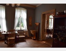 Ein wohnlicher Raum mit Holzboden, ausgestattet mit einem kleinen Holztisch und Stühlen vor zwei bodenlangen Fenstern mit weißen Vorhängen. Rechts steht ein Holzregal mit Gläsern und Geschirr, und durch eine Türöffnung ist ein Klavier in einem angrenzenden Raum sichtbar.