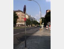 Straßenszene in einer Stadt mit einer mehrspurigen Fahrbahn, flankiert von Gehwegen und mehrstöckigen Wohngebäuden. Im Vordergrund sind eine Bushaltestelle, ein Fußweg sowie Straßenschilder zu sehen, während Bäume entlang der Straße Schatten spenden.