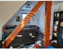 Zimmer unter einem Dach mit sichtbarem Holzträger, hellem Tageslicht und Möblierung. Im Raum stehen ein Schreibtisch mit Monitor, ein Bett, ein Regal mit Aktenordnern sowie eine Couch.