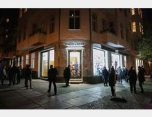 Straßenszene bei Nacht mit einer beleuchteten Buchhandlung „Einar & Bert Theaterbuchhandlung“ in einem Eckgebäude. Vor der Buchhandlung stehen mehrere Menschen in kleinen Gruppen auf dem Pflasterweg.