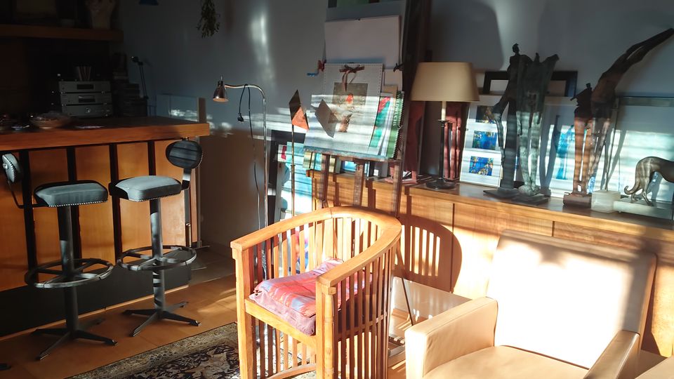 Ein gemütlicher Innenraum mit Holzboden und moderner Einrichtung, darunter ein hölzerner Stuhl, ein beiger Sessel und eine Theke mit Barhockern. Im Hintergrund stehen Kunstwerke, Staffeleien und Dekorationsobjekte, während Sonnenlicht durch ein Fenster den Raum beleuchtet.