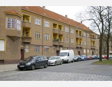 Wohnblock mit drei Stockwerken im typischen Stil der 1920er Jahre, gekennzeichnet durch erdfarbene Fassaden, gelb umrahmte Fenster und Balkone. Vor dem Gebäude parken Autos entlang einer mit Kopfsteinpflaster gesäumten Straße, daneben stehen vereinzelt Bäume.