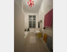 Schmales Badezimmer mit hoher Decke, zentralem Fenster und einem roten Kronleuchter an der Decke. Auf der linken Seite befindet sich eine Badewanne mit Pflanzen darauf, rechts ein Waschbecken, Handtücher an einem Heizkörper und ein Spiegel darüber.