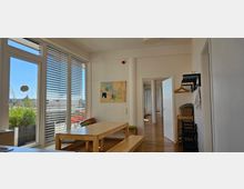 Heller Raum mit Holzboden, großen Fenstern mit Jalousien und Blick auf eine Terrasse mit Pflanzen. Im Vordergrund steht ein schlichter Holztisch mit zwei Bänken, an der Wand hängen ein bunter Plan und kleine Regale, daneben eine geöffnete Tür, die in einen Flur mit weißen Säulen führt.