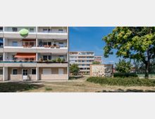 Wohnblock in einer gepflegten Wohnsiedlung mit grauer Fassade und Balkonen, die teilweise mit Blumen und Markisen dekoriert sind. Im Vordergrund ein grüner Bereich mit Bäumen, einer Hecke und einem Mosaik-Kunstwerk auf einer Steinwand.