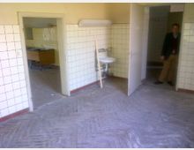 Ein leerer Raum mit weißen, gefliesten Wänden, einem kleinen Waschbecken und offenen Türen, die in andere Räume führen. Der Boden besteht aus abgenutztem Parkett, und es ist eine Person im Hintergrund sichtbar.