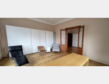 Heller Wohnraum mit Fischgrätparkett, hohen Decken und großem weißen Schrank an der Wand. Offene Holztüren mit Glasfeldern führen in einen angrenzenden Raum; davor stehen ein schwarzer Liegestuhl, ein kleiner Sessel und ein einfacher Tisch.