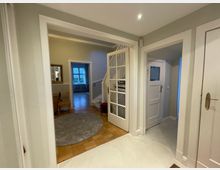 Ein Innenraum eines Hauses mit einem Flur, der in verschiedene Räume führt. Zu sehen sind eine geöffnete weiße Tür mit Glaseinsatz, eine Holztreppe, eine Wand mit einem Spiegel, ein Teppich auf Parkettboden und eine weitere geschlossene weiße Zimmertür.