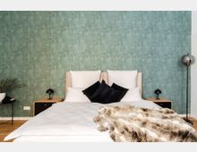 Schlafzimmer mit einem Doppelbett vor einer grünen Wand mit pflanzlichem Muster, weißen Kissen sowie schwarzen Dekokissen. Daneben stehen zwei Nachttische mit kleinen Lampen, im Vordergrund liegt eine gemusterte Kunstfell-Decke.