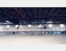 Große, verlassene Halle mit hohem Ziegeldach, unterstützt von Stahlträgern. Die Wände aus verputztem Mauerwerk sind stellenweise beschädigt und mit Graffiti bedeckt; wenige schmale Fenster lassen Licht herein.