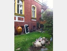 Backsteinfassade eines Gebäudes mit gelb umrahmten Fenstern, davor ein kleiner Garten mit Rasenfläche, einem Steinhaufen, einem großen Keramikgefäß und einem alten Verkehrssignal. Im Hintergrund sind Bäume, eine Holzabtrennung und Gartenaccessoires wie Pflanzentöpfe und ein Gartenschlauch zu sehen.