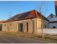 Modernes Wohnhaus mit Holzfassade und rotem Satteldach, das mit Solarpaneelen ausgestattet ist. Vor dem Gebäude stehen junge, blattlose Bäume entlang einer gepflasterten Gehwegkante in einer städtischen Straße bei Sonnenschein.