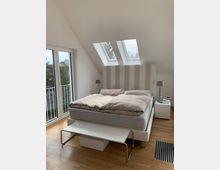 Ein modernes Schlafzimmer mit schräger Decke, zwei Dachfenstern und großen Glastüren, die zu einem Balkon führen. Ein großes Bett mit gepolstertem gestreiftem Kopfteil steht zentral, flankiert von Nachttischen mit Lampen; der Holzboden verstärkt den hellen und minimalistischen Eindruck des Raums.